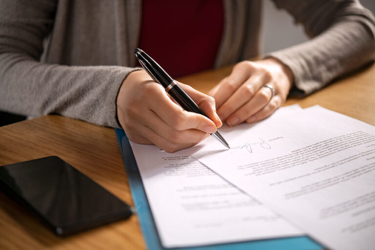 Close up of hands of young woman signing legal documents on desk. Close up of woman hand holding pen and signing legal paper seated at desk. Detail of lawyer filling official document, deal done.
